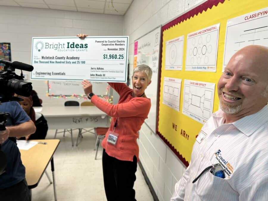 Two people joyfully present a large grant check in a classroom while a camera records the event for documentation.