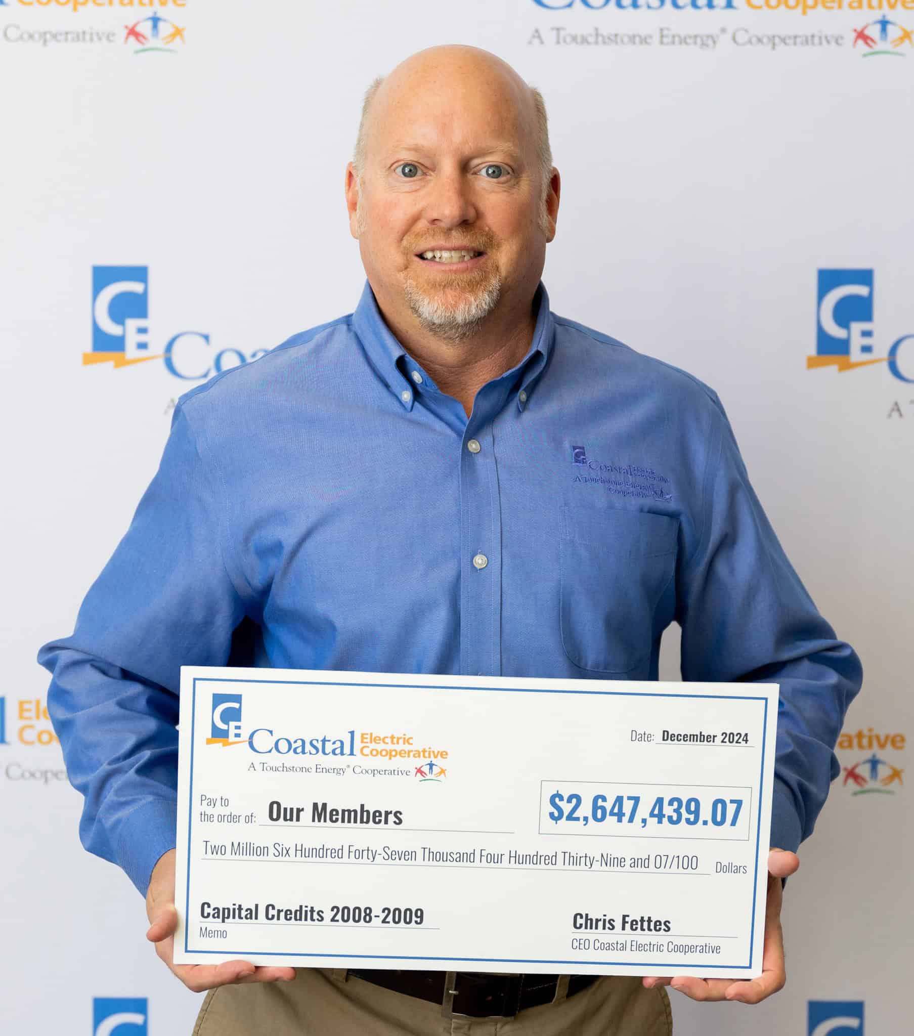 A person in a blue shirt holds a large check from Coastal Electric Cooperative for over $2.6 million, dated December 2024.