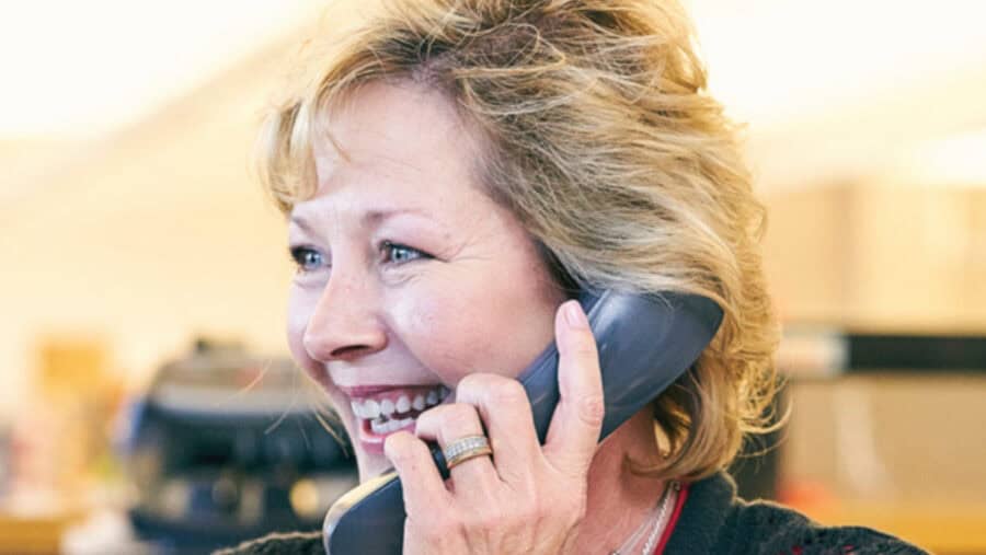 A person smiling while talking on a corded telephone, with a blurred background suggesting an office environment. No recognizable landmarks or historical buildings.