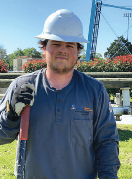 A person in a hard hat and gloves stands outdoors on grass, holding a tool. Trees and equipment are visible in the background.