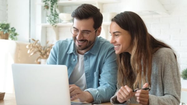 Two people smiling while looking at a laptop in a bright, modern home office setting with plants and shelves in the background.