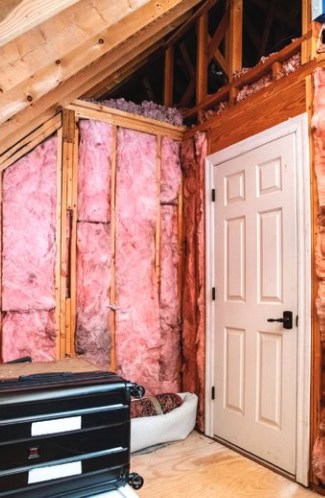 An attic under construction, showing exposed insulation and wooden framework. A white door is closed, and a black suitcase is nearby.