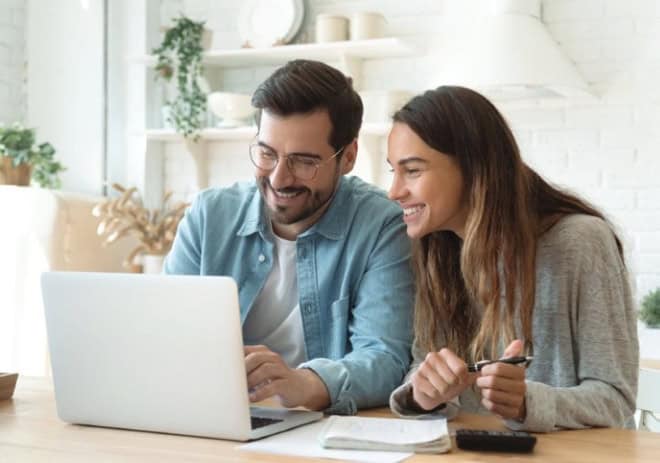 Two people smiling and working on a laptop in a bright, cozy kitchen with greenery and modern decor on shelves in the background.