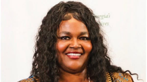 A person with long curly hair smiles at the camera against a plain background. The person is wearing a decorative top.