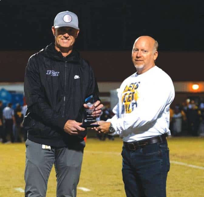 Two people in casual attire stand on a sports field, one holding a trophy. Nighttime setting with indistinct background activity.