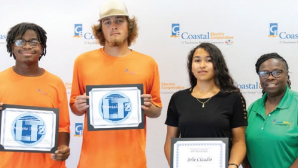 Four people standing with certificates in front of a Coastal Electric Cooperative backdrop, smiling at the camera. No recognizable landmarks or historical buildings.