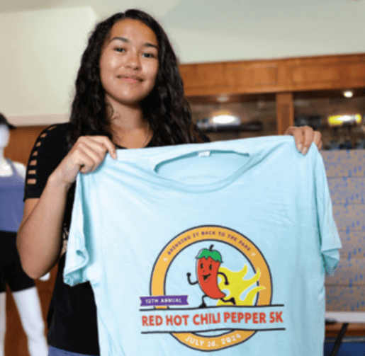 A person holds a "Red Hot Chili Pepper 5K" shirt inside a shop. A mannequin is visible in the background.