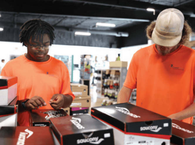 Two people in orange shirts handle shoe boxes inside a store. The interior displays various products on shelves in the background.