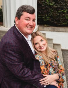 Two people smiling, sitting on outdoor steps with greenery in the background. The person on the left is wearing a jacket, and the other has patterned clothing.